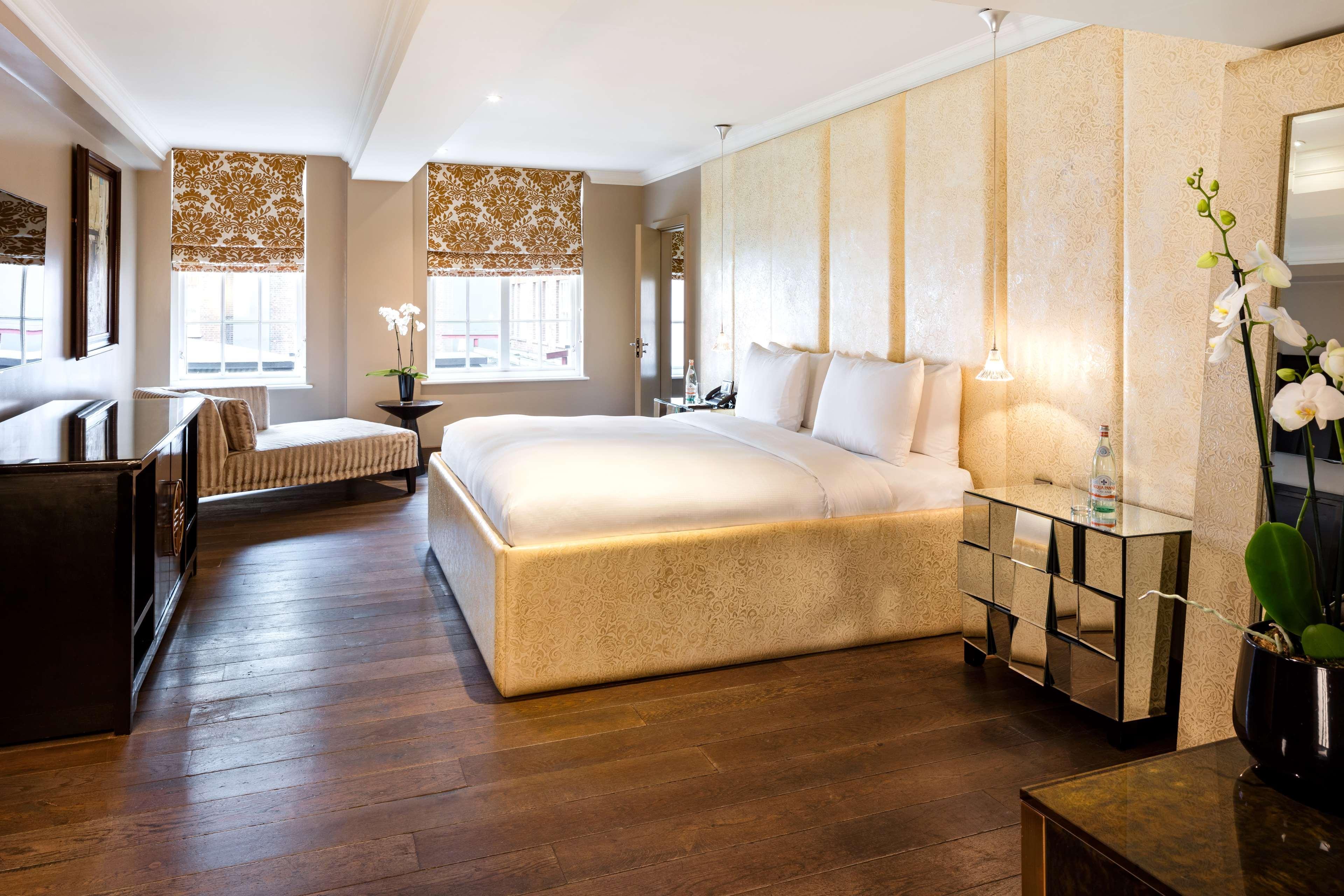 The May Fair, A Radisson Collection Hotel, Mayfair Londýn Exteriér fotografie The photo shows a modern and elegantly designed hotel room or bedroom. It features a large, plush bed with two pillows, situated against a light-colored wall. To one side, there is a stylish bench or chaise lounge, adding a comfortable seating area. 