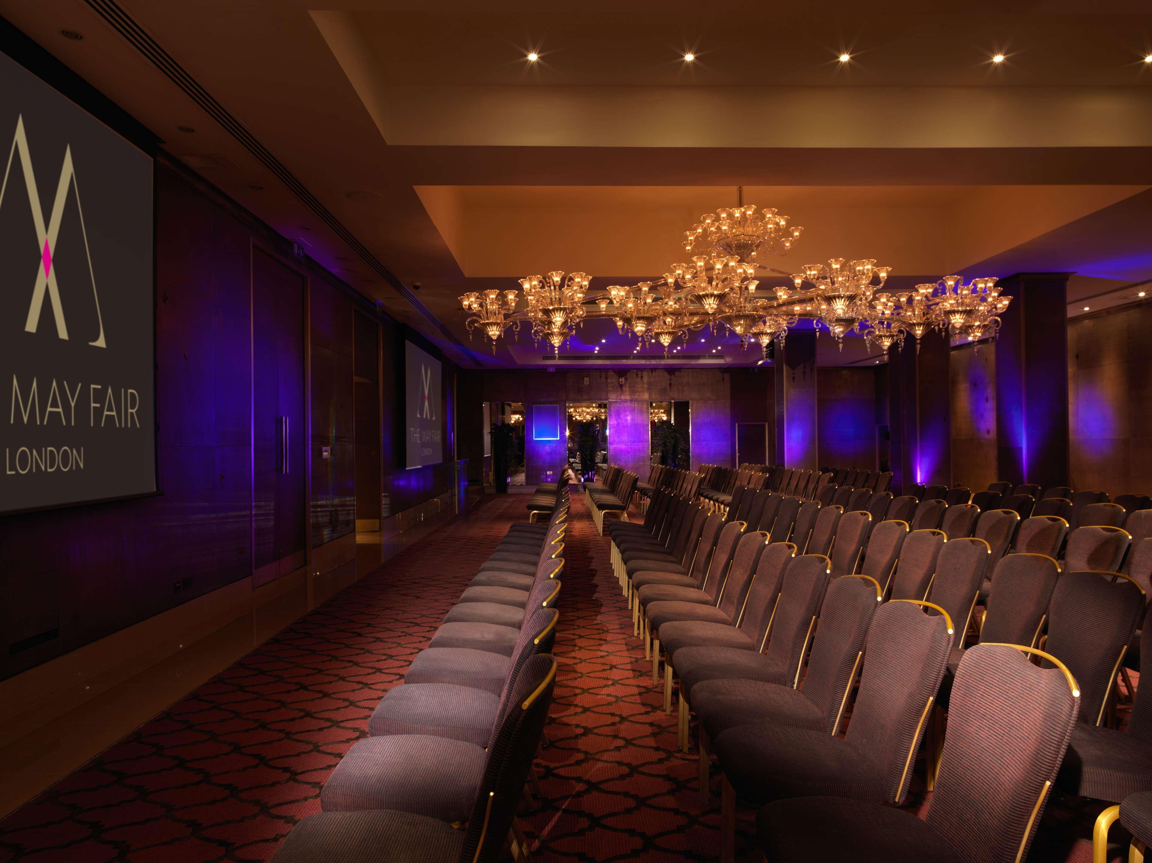 The May Fair, A Radisson Collection Hotel, Mayfair Londýn Exteriér fotografie The image shows a conference or event space with rows of elegant, upholstered chairs facing a large screen. The room is adorned with chandeliers that provide ambient lighting, and there are purple lighting accents around the space. The branding for "