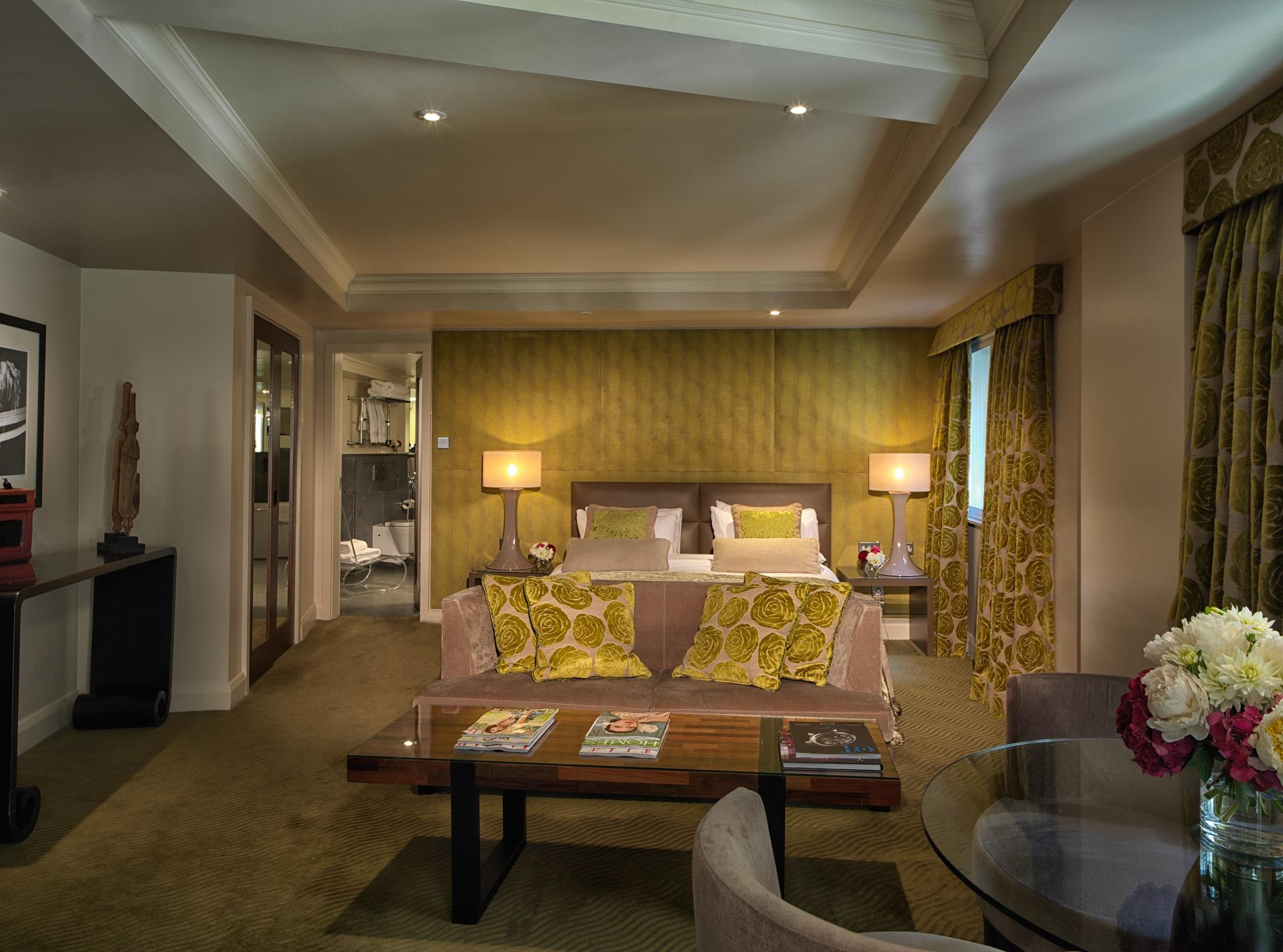 The May Fair, A Radisson Collection Hotel, Mayfair Londýn Exteriér fotografie The photo showcases a stylish hotel room or suite. It features a seating area with a couch adorned with decorative yellow pillows and a coffee table. There are two lamps on either side of the couch providing soft lighting. The walls are painted in a 