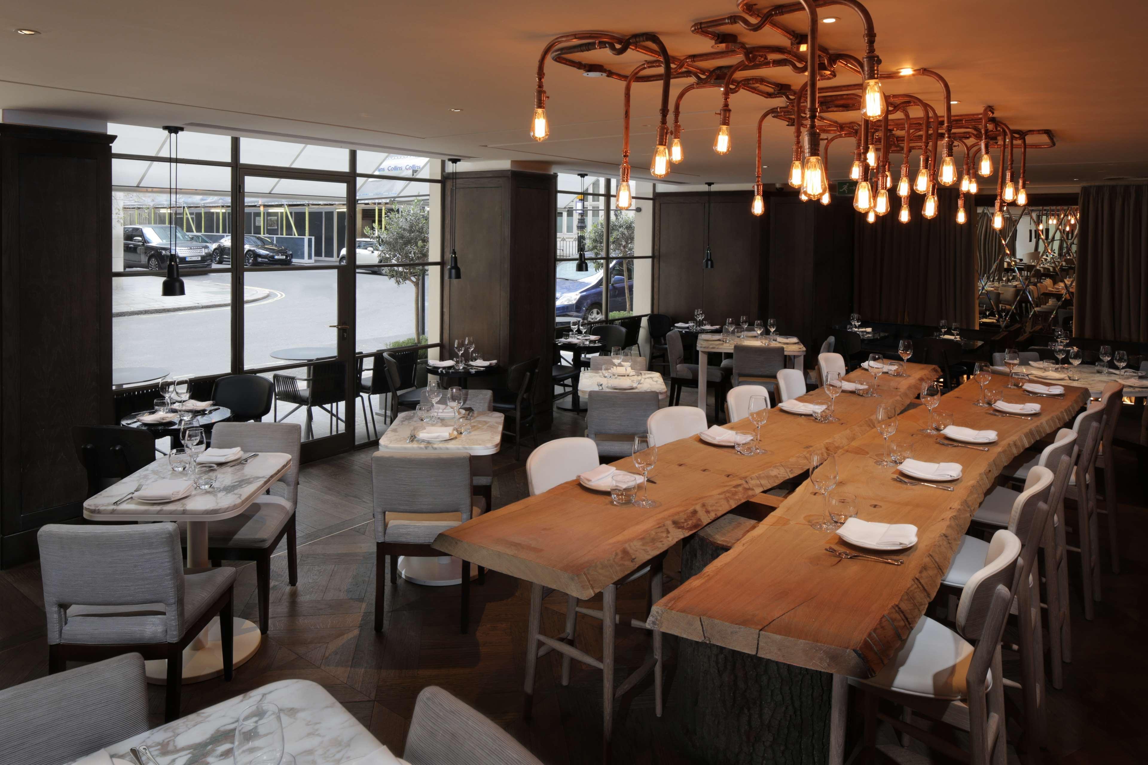 The May Fair, A Radisson Collection Hotel, Mayfair Londýn Exteriér fotografie The photo displays a stylish restaurant interior featuring a mix of wooden and upholstered furniture. There's a large wooden table at the center, surrounded by various seating options, including chairs and smaller tables. The room is well-lit, with u
