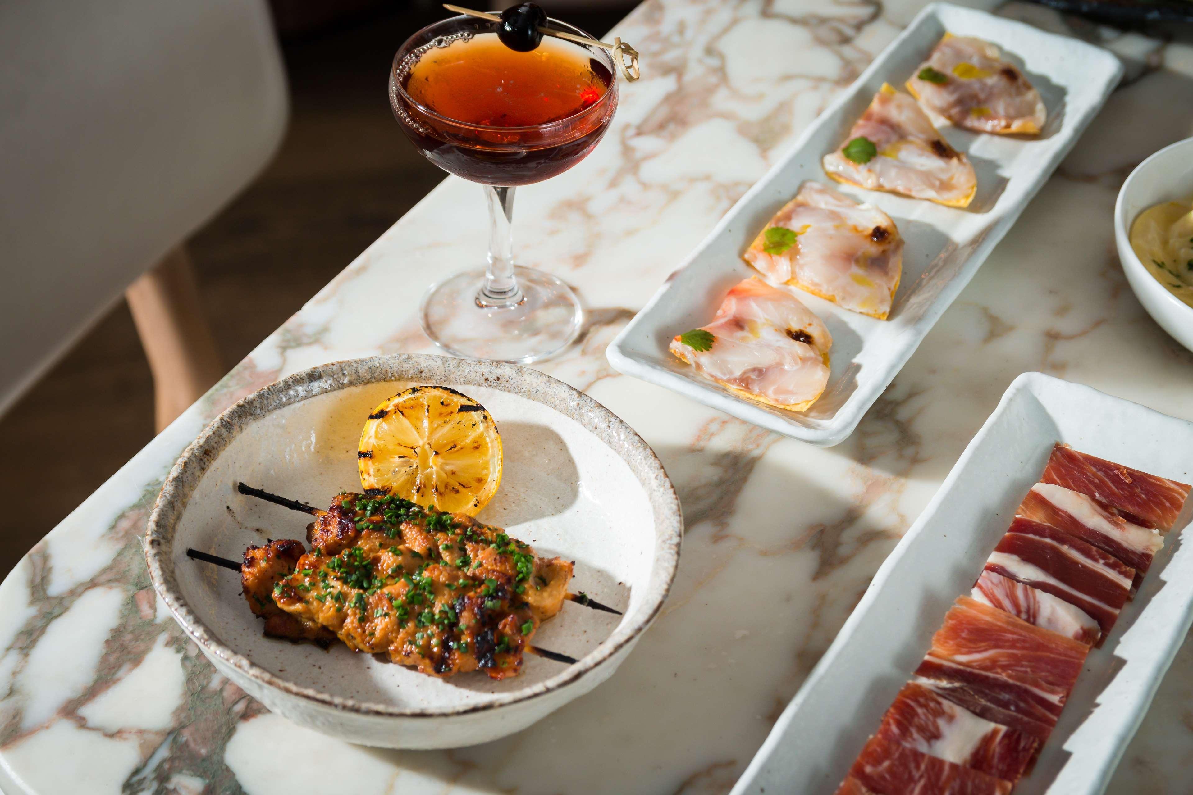 The May Fair, A Radisson Collection Hotel, Mayfair Londýn Exteriér fotografie The photo features a beautifully arranged dining table with a variety of food and a drink. In the foreground, there is a dish containing grilled or cooked meat, garnished with herbs and served with a roasted lemon slice. Next to it, a cocktail drink 