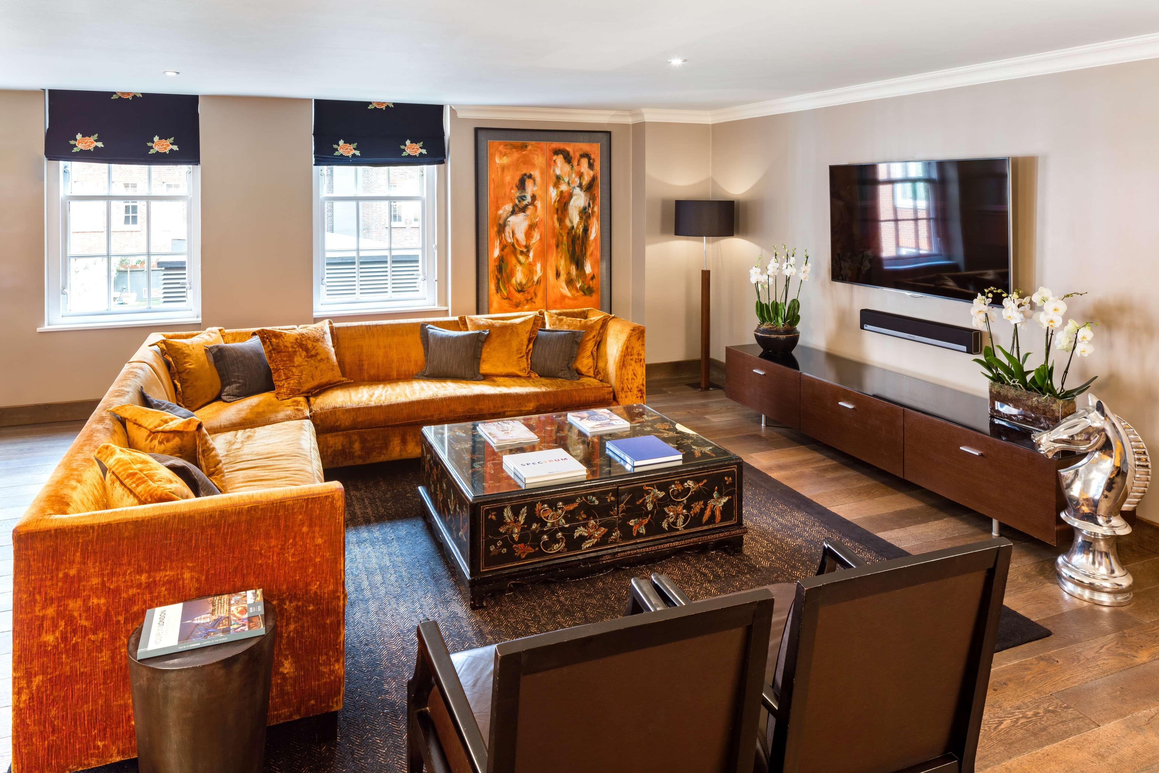 The May Fair, A Radisson Collection Hotel, Mayfair Londýn Exteriér fotografie The photo shows a modern living room with a stylish design. It features a large, orange sectional sofa with colorful cushions, positioned around a coffee table that has an ornate design. The floor is wooden, and there's a dark area rug beneath the se