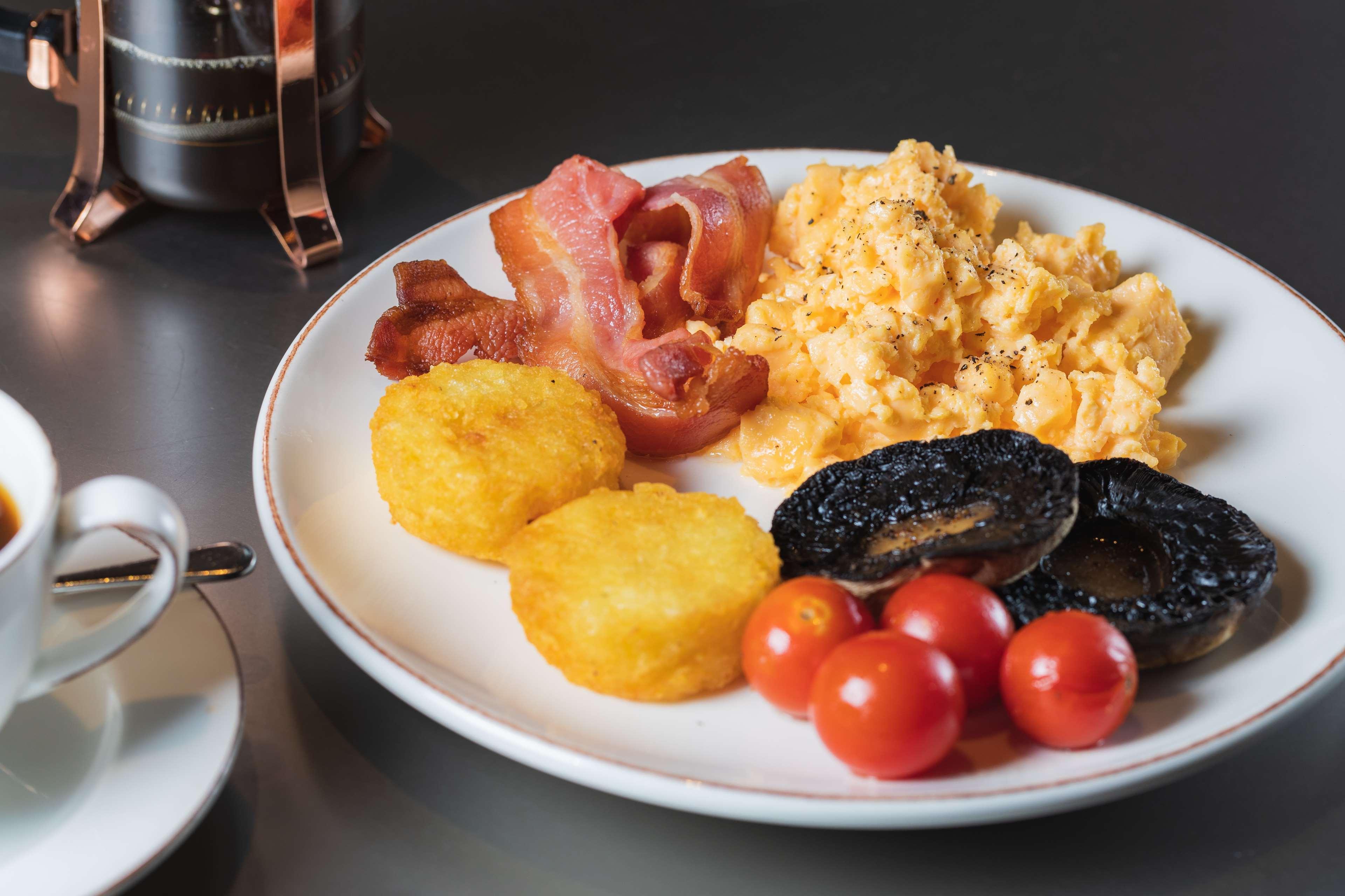 The May Fair, A Radisson Collection Hotel, Mayfair Londýn Exteriér fotografie The photo shows a plate of breakfast food that includes scrambled eggs, crispy bacon strips, two golden hash browns, grilled tomatoes, and slices of black pudding. The dish appears to be served alongside a small cup, presumably for coffee or tea. The