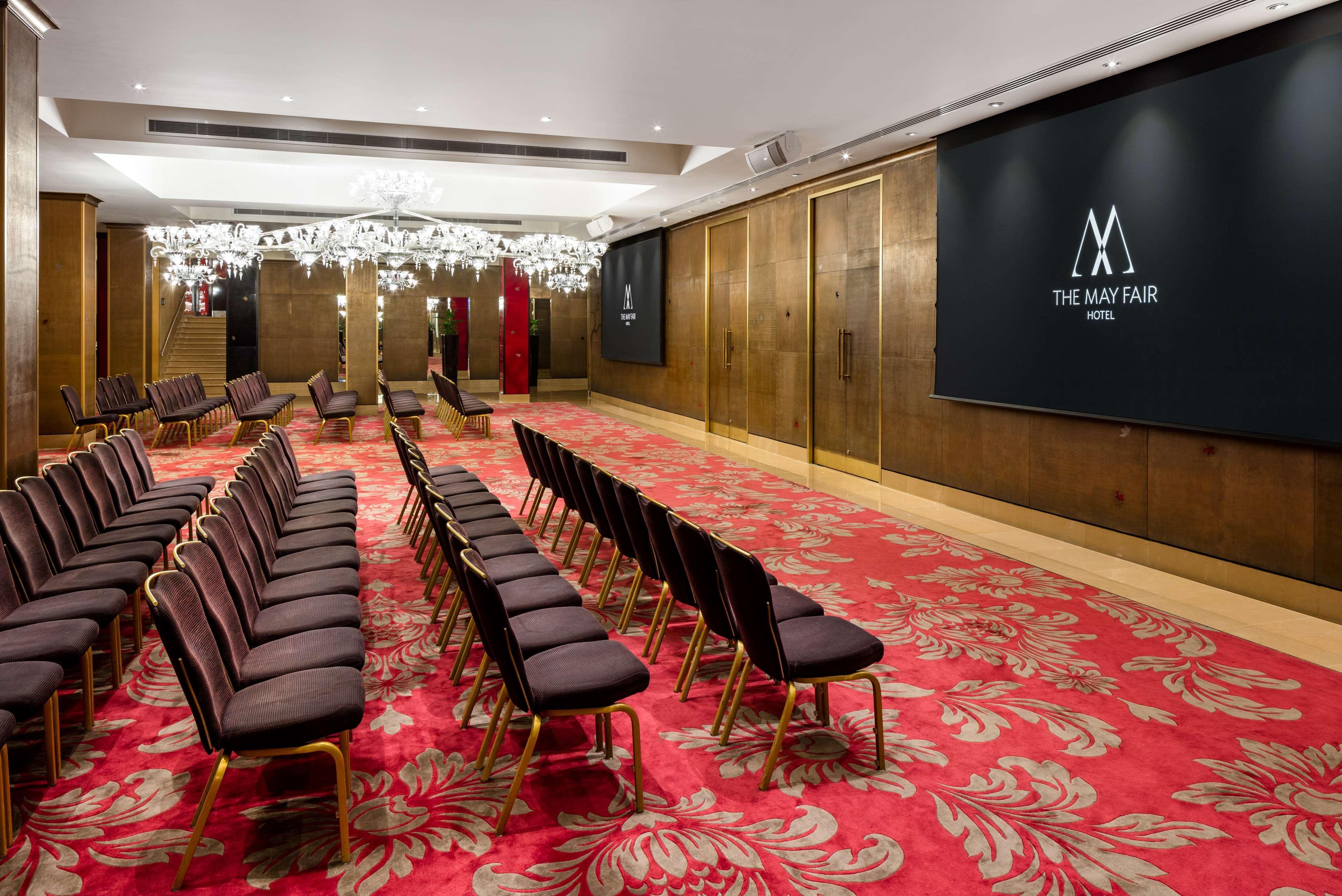 The May Fair, A Radisson Collection Hotel, Mayfair Londýn Exteriér fotografie The photo shows a conference or event room inside The May Fair Hotel. The space has a stylish design with decorative chandeliers hanging from the ceiling. There are rows of upholstered seating arranged in front of a large screen that displays the hot