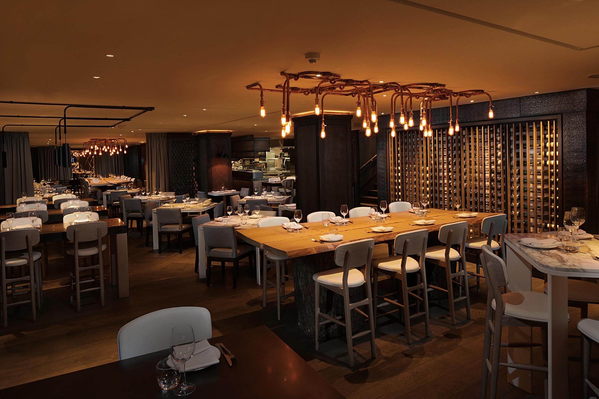 The May Fair, A Radisson Collection Hotel, Mayfair Londýn Exteriér fotografie The photo depicts a modern restaurant interior. The space features a combination of wooden and white furniture, with tables set for dining. There is a long, communal-style table at the center, surrounded by high chairs. The lighting is warm, coming f