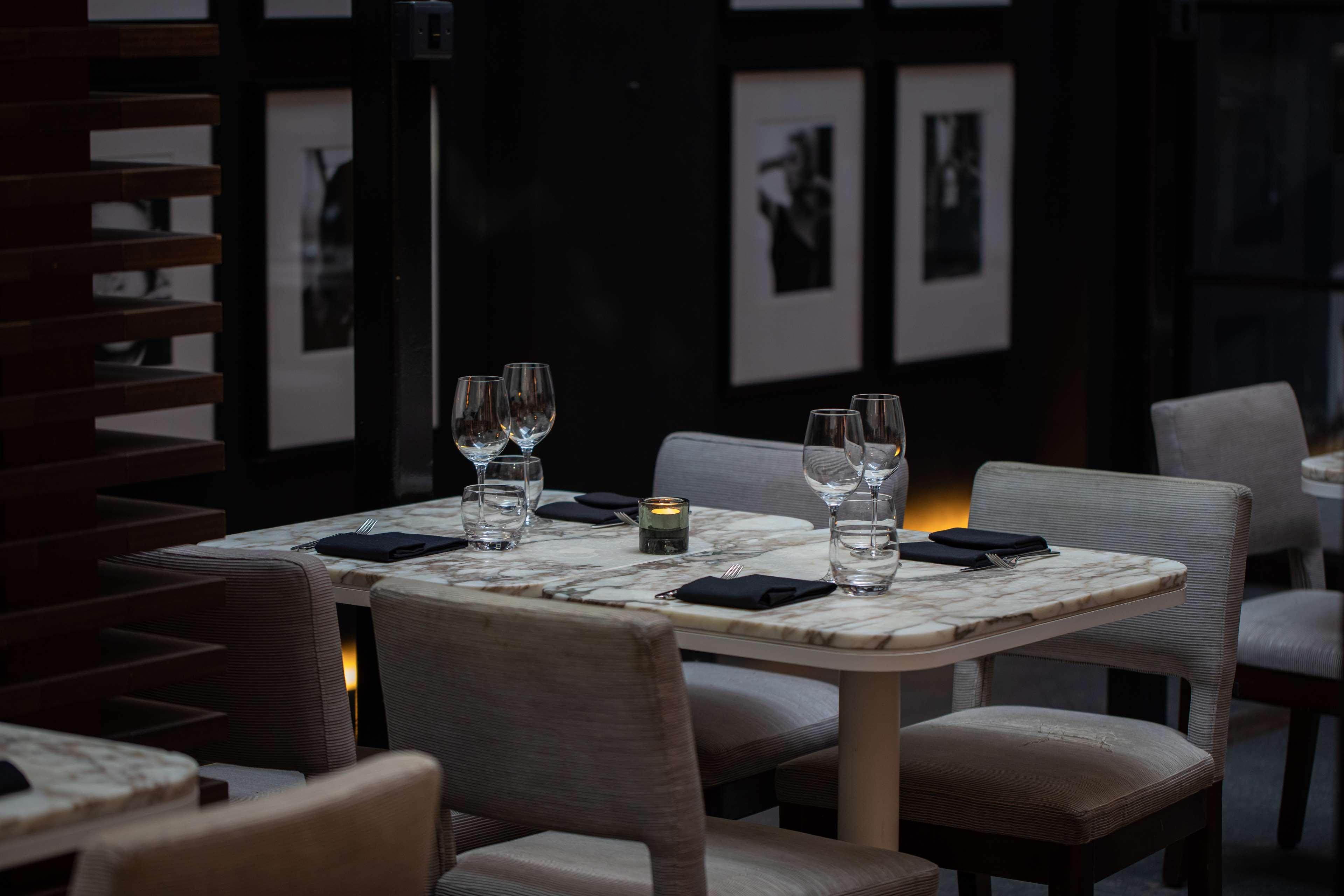 The May Fair, A Radisson Collection Hotel, Mayfair Londýn Exteriér fotografie The photo shows a dining area with a stylish and elegant atmosphere. There are two tables set for dining, featuring marble tops and light-colored chairs. Each table has wine glasses and a small candle holder, creating a warm ambiance. In the backgrou