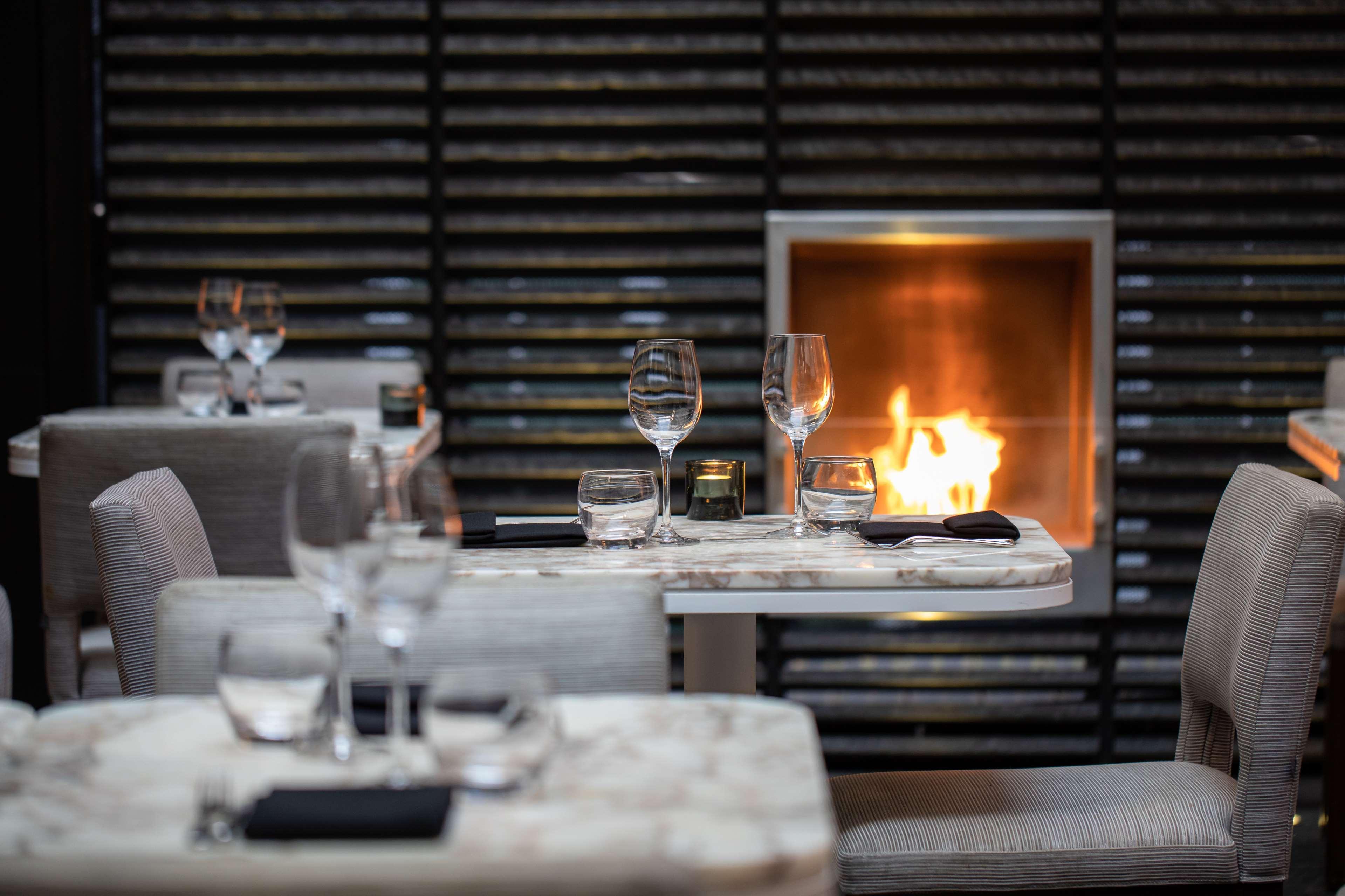 The May Fair, A Radisson Collection Hotel, Mayfair Londýn Exteriér fotografie The photo depicts an elegant dining space featuring tables with marble tops, each set with wine glasses. The chairs are upholstered in a soft, neutral fabric. In the background, there is a modern fireplace that adds warmth to the atmosphere with visi