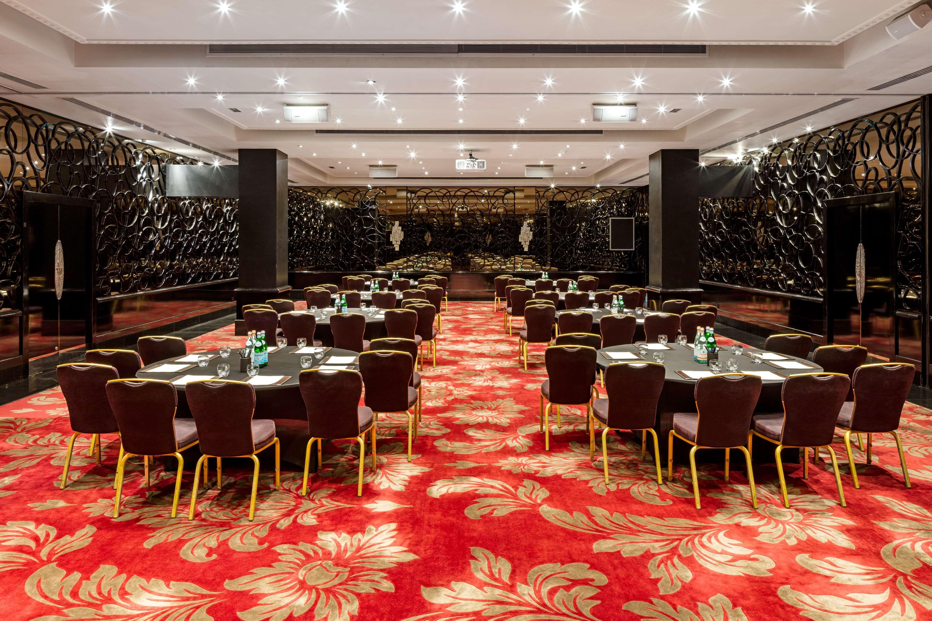 The May Fair, A Radisson Collection Hotel, Mayfair Londýn Exteriér fotografie The photo shows a spacious dining or banquet room. It features several round tables arranged neatly with dark chairs. The floor is covered with a vibrant red carpet that has floral patterns. The walls are decorated with a sleek, dark design, adding a