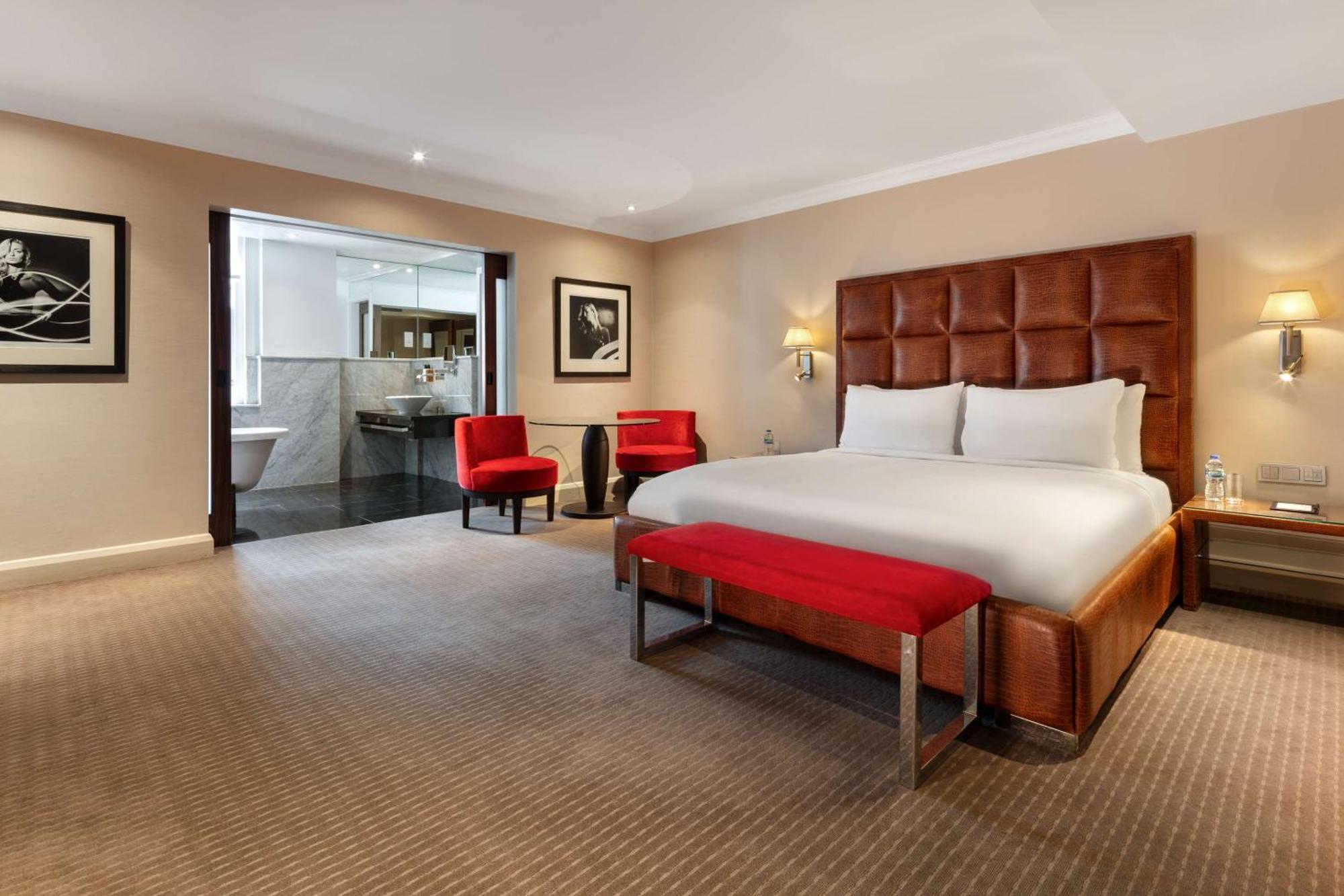 The May Fair, A Radisson Collection Hotel, Mayfair Londýn Exteriér fotografie The photo shows a modern hotel room featuring a large, comfortable king-size bed with a beige and brown color scheme. There are two red accent chairs positioned near the bed, adding a pop of color. A small red bench is placed at the foot of the bed. 