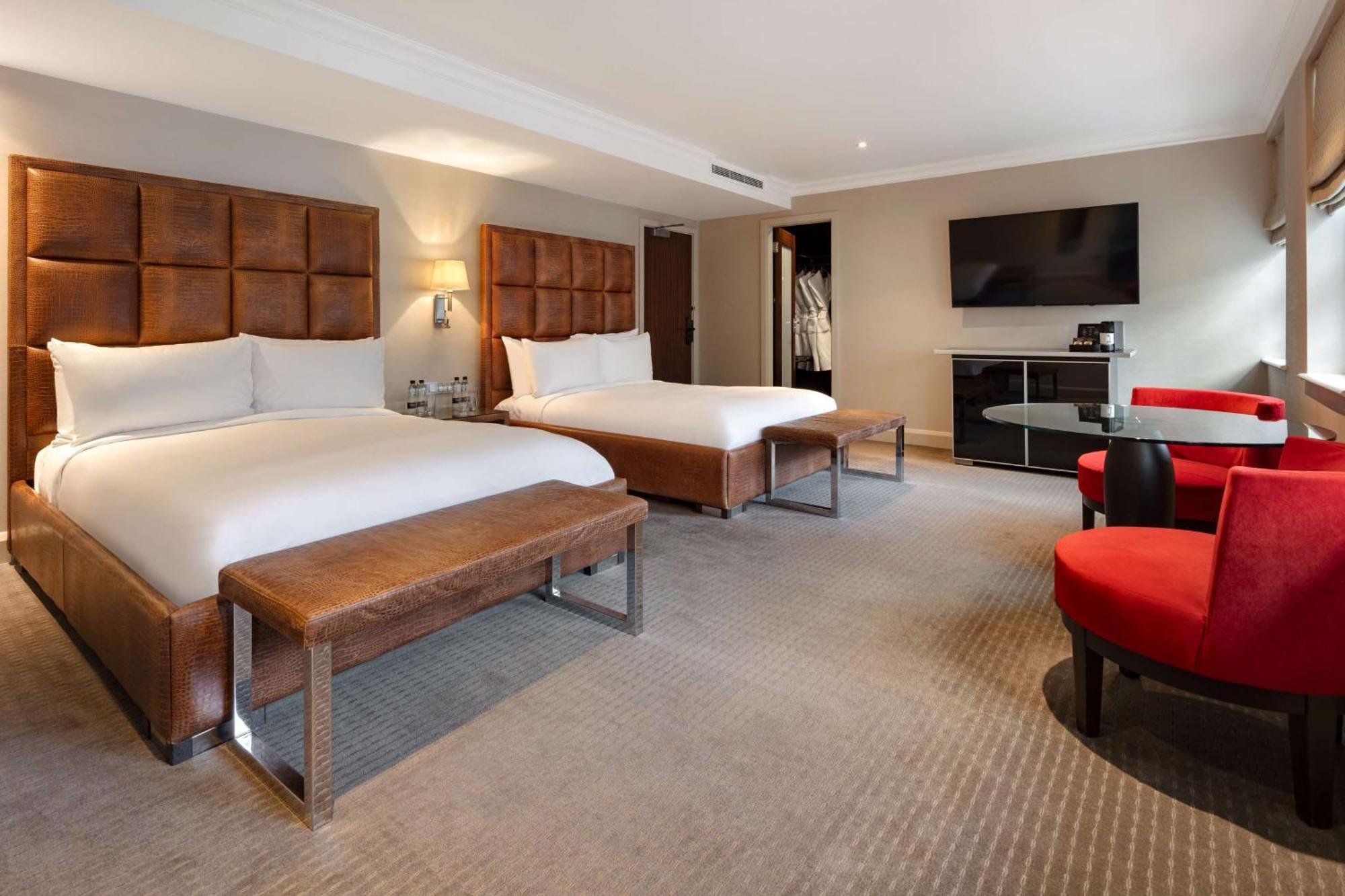 The May Fair, A Radisson Collection Hotel, Mayfair Londýn Exteriér fotografie The photo shows a modern hotel room with two beds. Each bed is made with white linens and has a large, upholstered headboard. There is a bench at the foot of each bed. To the right, there are two red chairs around a small glass table. A flat-screen T