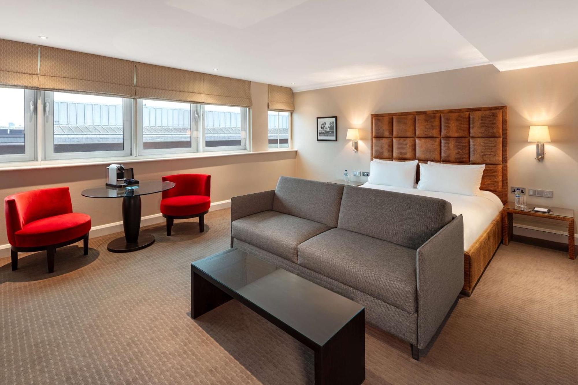The May Fair, A Radisson Collection Hotel, Mayfair Londýn Exteriér fotografie The photo shows a modern hotel room featuring a comfortable setup. There is a large double bed with a headboard, some decorative artwork on the wall, and two windows allowing natural light to enter. In the room, there's a gray sofa facing a dark coff