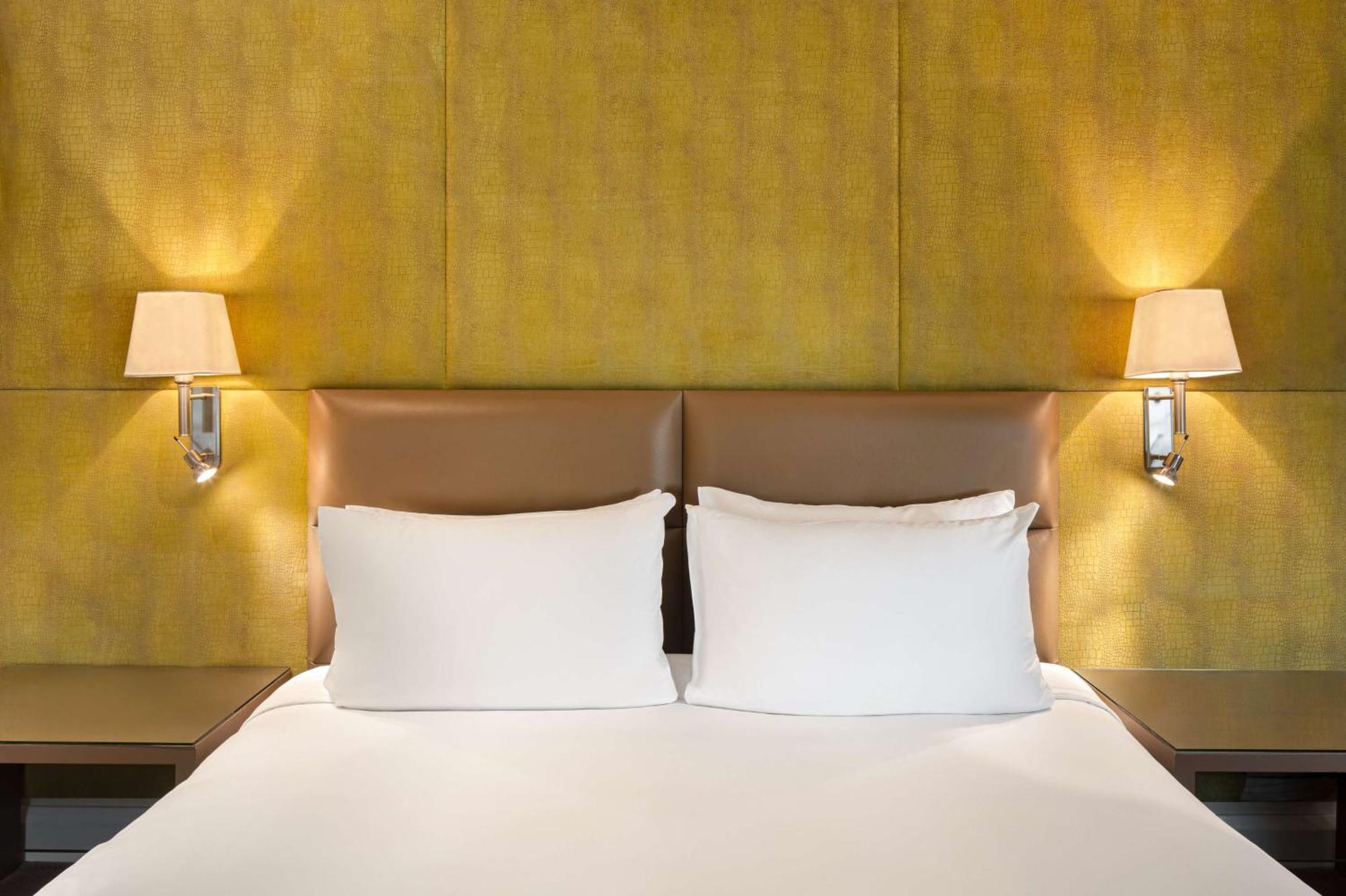 The May Fair, A Radisson Collection Hotel, Mayfair Londýn Exteriér fotografie The photo shows a cozy bedroom setting featuring a large bed with two white pillows. The headboard has a smooth, light brown leather material, and there are two wall-mounted lamps on either side of the bed, illuminating a textured yellow wall behind 