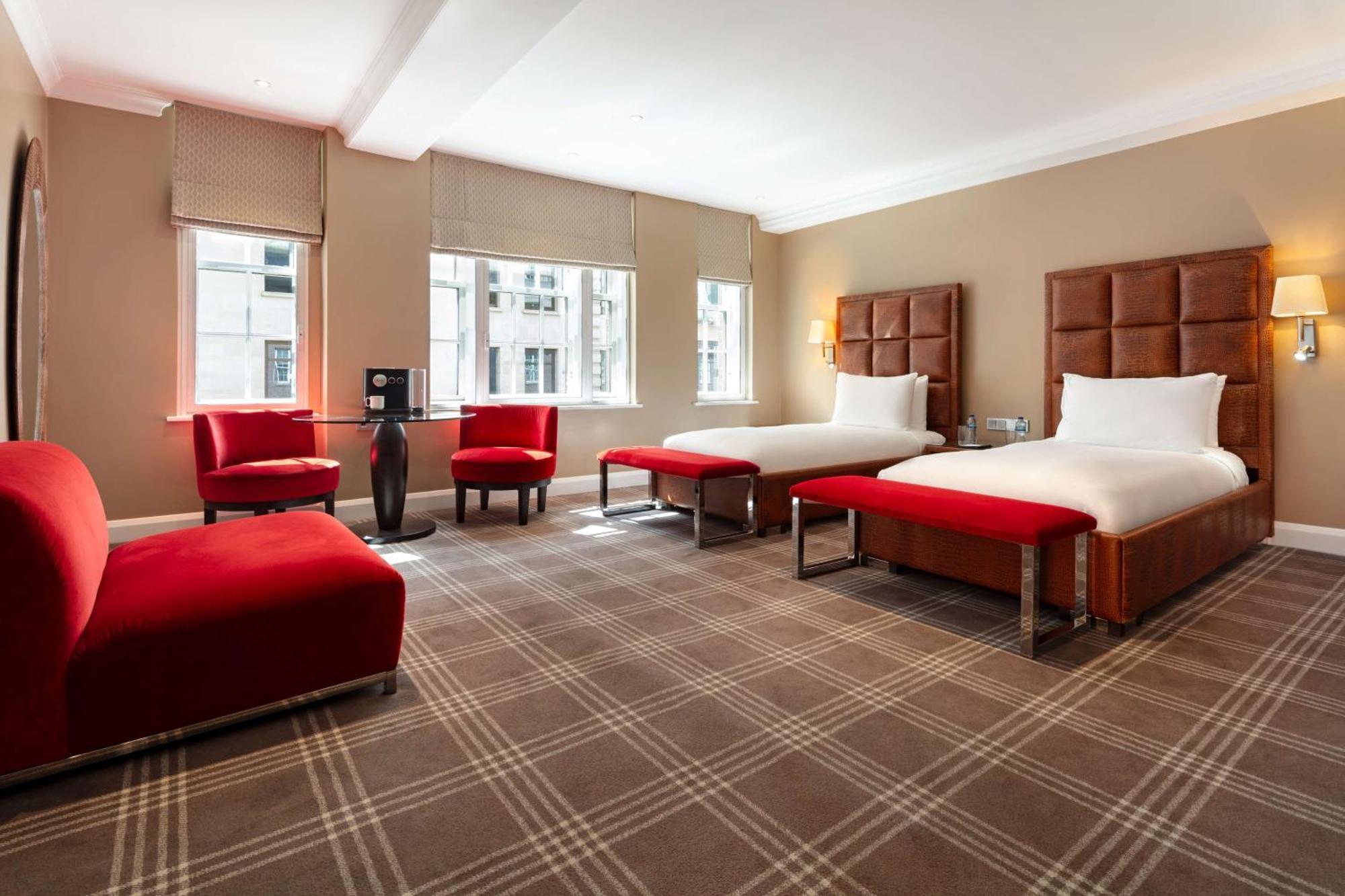 The May Fair, A Radisson Collection Hotel, Mayfair Londýn Exteriér fotografie The photo shows a modern hotel room featuring two beds with white bedding and brown headboards. There are two red benches at the foot of each bed. A red upholstered chair and a small round table are positioned near the window, which allows natural li