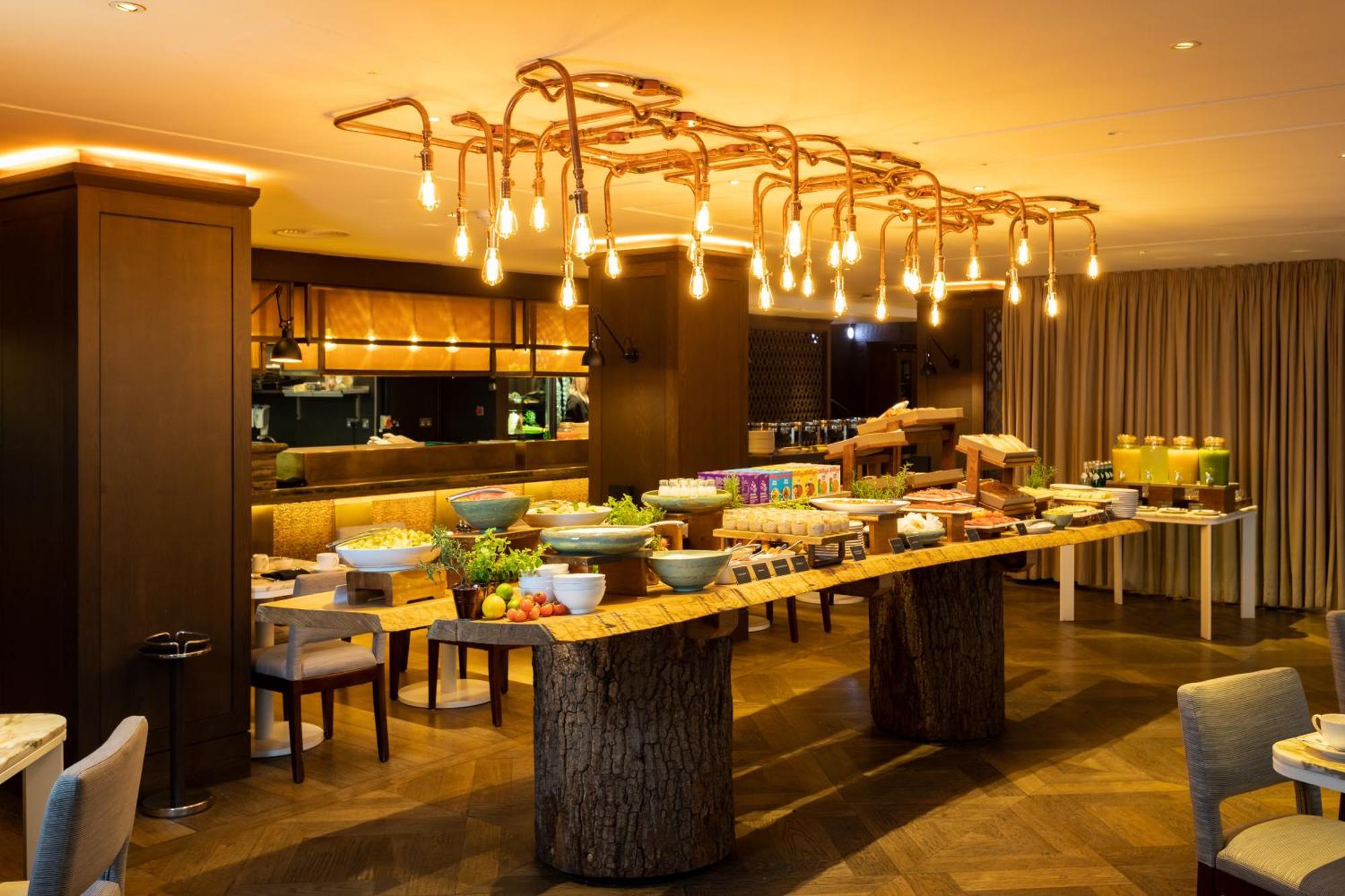 The May Fair, A Radisson Collection Hotel, Mayfair Londýn Exteriér fotografie The photo shows a stylish dining area with a buffet setup. In the center, there is a long wooden table made from a tree trunk, displaying various dishes and food items. The setting features an elegant lighting fixture made of exposed bulbs that resem
