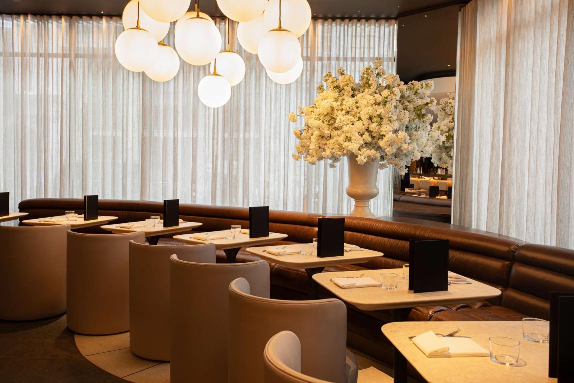 The May Fair, A Radisson Collection Hotel, Mayfair Londýn Exteriér fotografie The photo shows a modern restaurant interior. It features stylish seating arrangements with curved booths and elegant tables. The lighting is soft, provided by large, round pendant lights hanging from the ceiling. A large floral arrangement in a whit